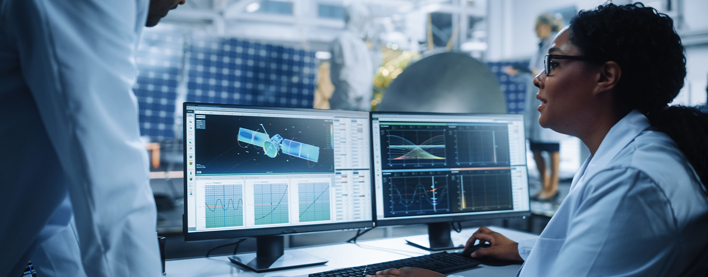 Female systems engineer sitting at computer looking at satelite schematics with another employee